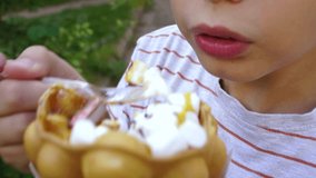 Closeup of mouth of white kid eating tasty sweets outdoors in street. Child holds big waffle cone filled with ice-cream, marshmallow and other sweets. Focus at lips. Real time 4k video footage.