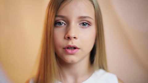 Happy face little girl with a make-up looks at camera talk speak stand indoors smile cute young attractive blonde hair beautiful caucasian female pretty close up portrait slow motion