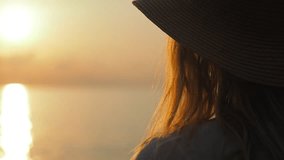 Young woman in a straw hat relaxing, looking at sea during sunset. Lifestyle video, people in nature concept. 