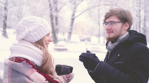 Young millennial woman and bearded man with glasses talking, smiling and drinking hot cocoa from cups in hands in winter park. 4K slow-motion footage.