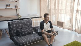 Pleasant-looking young unemployed Caucasian man sitting on sofa and playing video game. Disgruntled shocked girlfriend looking at lazy boyfriend. Problems in relationship.