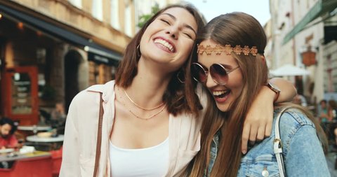 Close up of two cheerful stylish and friendly Caucasian girls walking the city street full of cafes and laughing together. Outside.