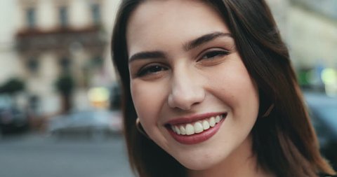 Close up of the cheerful charming young Caucasian woman with short dark hair smiling to the camera in the city street. Portrait. Outdoors.