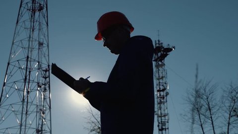 Silhouette of an engineer at sunset, who looks at the telecommunication pillars. Engineer writes data to the tablet. The concept of modern telecommunications