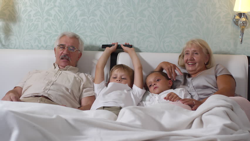 Cute little boy and girl lying on cozy bed between grandmother and grandfather while watching TV together at home | Shutterstock HD Video #1019360974