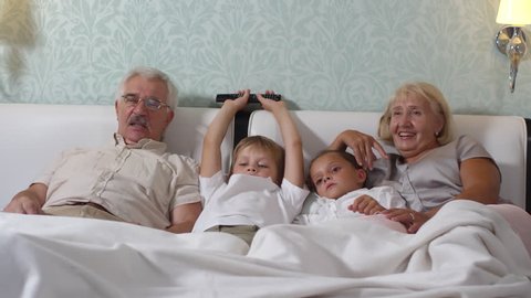 Cute little boy and girl lying on cozy bed between grandmother and grandfather while watching TV together at home
