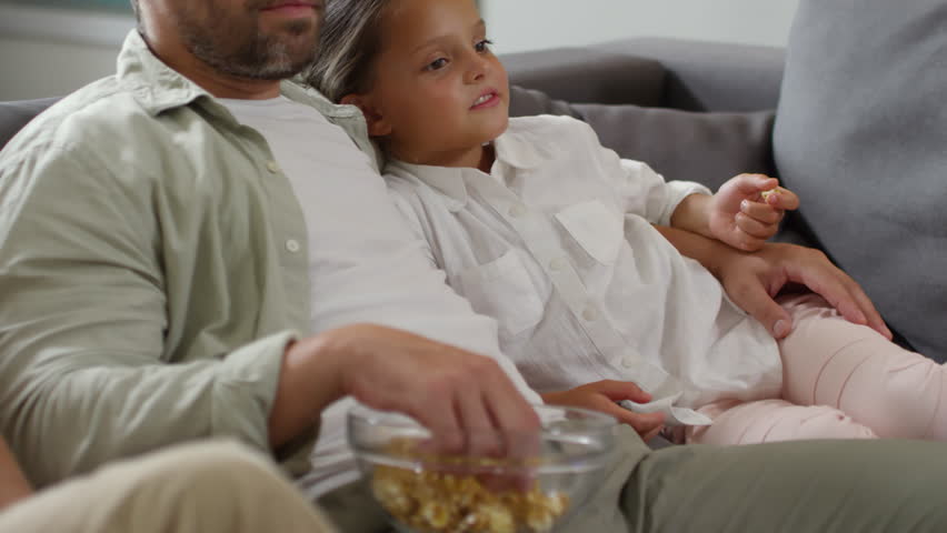 Bearded father and little daughter embracing and eating popcorn while sitting together on sofa and watching TV
