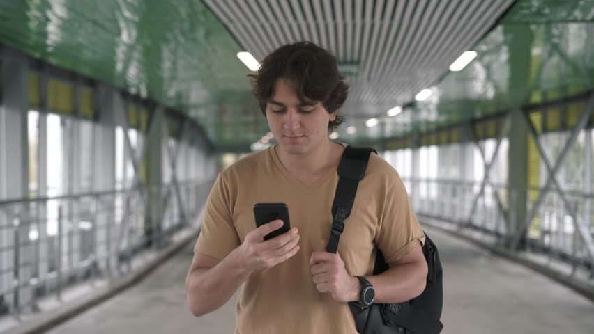 Attractive young caucasian man wearing beige t shirt and backpack is walking in tunnel, smiling and talking on smartphone. Tracking real time medium shot