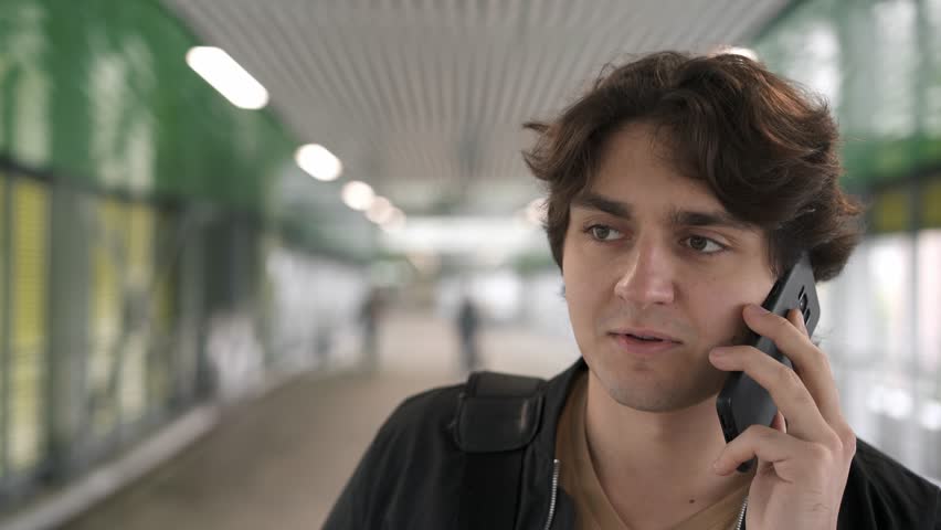 Handsome young man with dark hair wearing black jacket is standing in tunnel and talking on his phone. Handheld real time close up shot
