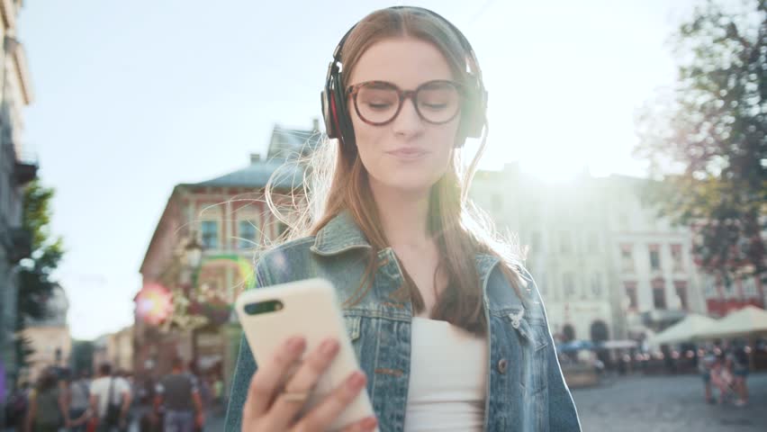 Smiling young woman with glasses listening to music in headphone use smartphone at city walk sunset look around close up portrait slow motion