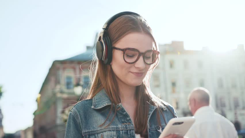 Attractive young woman with glasses listening to music in headphone use smartphone at city walk sunset look around smile portrait close up slow motion | Shutterstock HD Video #1019386018