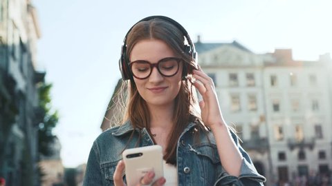 Attractive young woman with glasses listening to music in headphone use smartphone at city walk sunset look around smile portrait close up slow motion