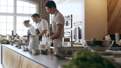 Pan shot of focused professional chefs cooking on a large counter top full of pots and tools in a kitchen with soft day lighting. Medium shot on 4k RED camera.