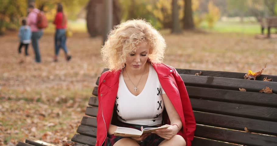 Pretty young woman reading a book on the bench outdoors at sunset.