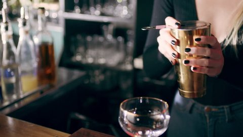 Pan shot of young female mixologist using strainer to pour a drink into a cocktail glass on a counter in a fancy bar with soft interior lighting. Medium shot on 4k RED camera.