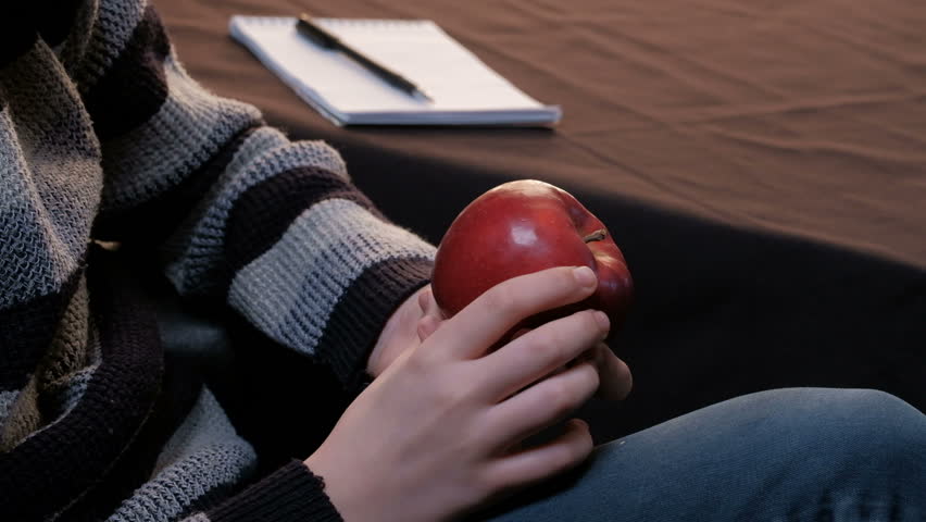 The guy is considering a ripe red apple. Closeup of the hands of a young man in a gray sweater. Student, artist or writer. Notebook and pen lie on the table in the background.