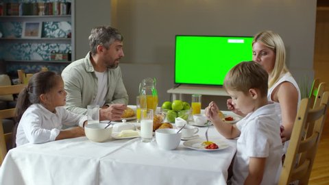 Beautiful Caucasian family having breakfast in dining room: mother and dad eating fried eggs and croissants, talking with son and daughter and watching TV with chroma key screen
