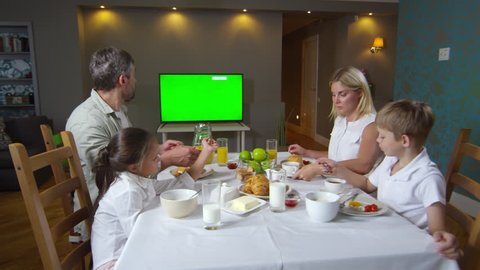 Panning shot of beautiful parents talking with little daughter and son while having breakfast together and watching TV with chroma key green screen
