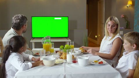Caucasian family having breakfast in the kitchen: mother and father eating fried eggs and croissants, talking with children and watching TV with green chroma key screen