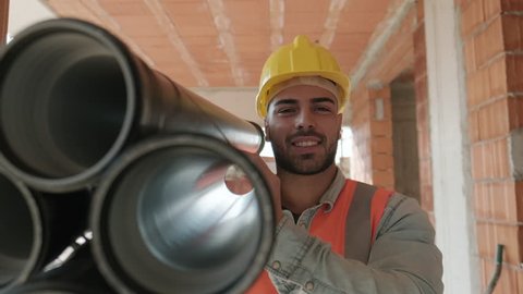 Proud people working in construction site. Portrait of happy white man at work in new house inside apartment building. Professional latino worker carrying pipes and smiling at camera. Slow motion