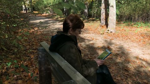 Senior woman holds her tablet sitting on a bench in a forest in autumn