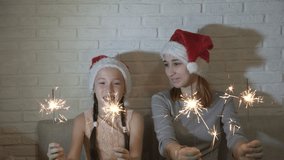 Cheerful, happy children in Santa's hats hold lighted sparklers in their hands, wave them and smile sitting on a sofa against a white brick wall. Close up. 4K. 25 fps.