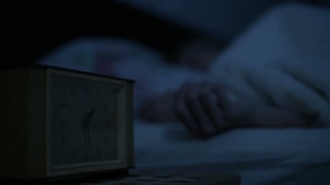 Close-up of a girl in pajamas lying in bed and unable to sleep looking at an alarm clock. Variable focus from hours to face girls. The effect of the American night. Low key cold light imitation night