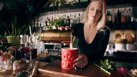 Camera moves towards a serious female bartender preparing a cocktail in a tiki glass then adding ice in interior fancy bar with soft day lighting. Medium to close up shot on 4k RED camera.