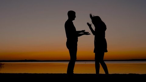 At sunset on the beach, silhouette of a man quarrels with a woman, she slaps him, beats with hands and swears, pushes him away, he begs her to forgive him, she go away. Slow motion.