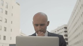 Thoughtful businessman working with laptop near office building. Handsome Caucasian manager thinking and using digital device outdoors. Remote work concept