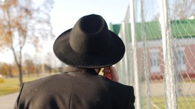 Man in a black stylish suit and hat walks along a grid fence and talks on the phone. Solving problems, business issues and tasks. View from the back. Lawyer. Mafia member. Slow motion handheld shot