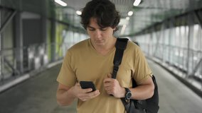 Handsome young man with dark hair wearing a beige t shirt and holding a backpack walking and web surfing on his smartphone. Tracking slow motion medium shot - Powered by Shutterstock - Get 15% off with code: PIKWIZARD15