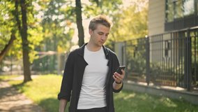 Side view of young man wearing black jacket walking in the park and texting with his smartphone Tracking slow motion medium shot