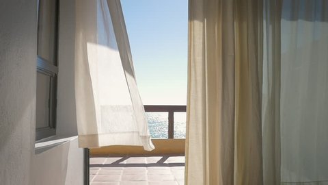 White curtains on the porch of a oceanfront resort blowing from a summer breeze in slow motion. Inside looking out windows framed by linen drapes onto the veranda of a luxury hotel with an ocean view.