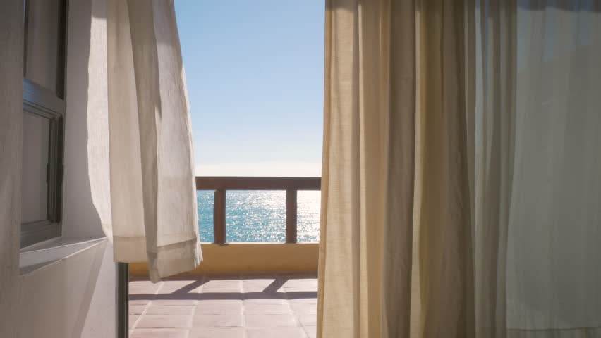 Balcony of a tropical luxury hotel by the ocean in 4k. Inside looking out at white curtains blowing in a warm summer wind with the ocean stretching out in the background. Wide shot depicts wanderlust. Royalty-Free Stock Footage #1019453176