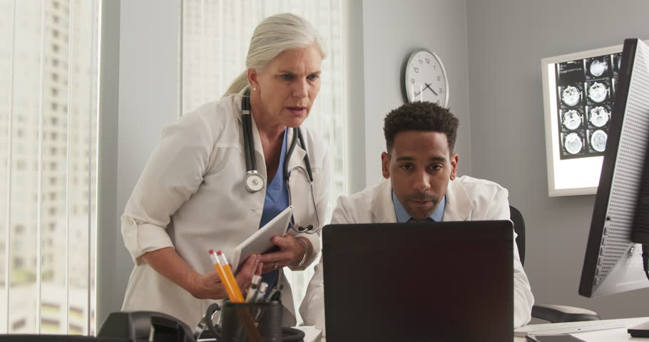 Two medical doctors working inside office and using laptop computer. Young black physician working on notebook computer while consulting senior colleague | Shutterstock HD Video #1019454991