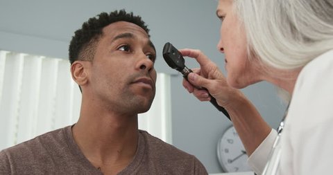Young black male patient having his eyes checked by senior female doctor. Mature female doctor using ophthalmoscope to examine eyes of african-american patient