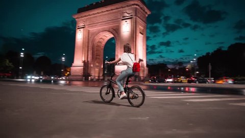 Tracking shot young woman biking riding a bike in front of Arch of Triumph Bucharest at dusk sunset