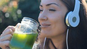 Beautiful woman listening to music with headphones and drinking green smoothies outdoor on a sunny summer day.