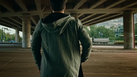 Backshot of an Athletic Young Man Walking Towards the Road Under a Bridge in an Urban Environment. He's Wearing a Grey Hoodie and a Sports Bag.