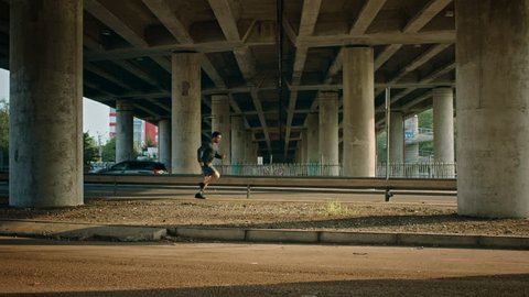 Athletic Muscular Man in Sports Outfit is Energetically Running in the Street. He is Jogging in an Urban Environment Under a Brindge with Cars in the Background.