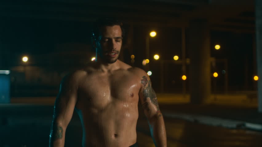 Shirtless Athletic Muscular Young Man is Celebrating His Sport Accomplishments on a Rainy Night. He is in an Urban Environment Under a Brindge with Cars in the Background.
