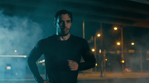 Sweating Athletic Muscular Young Man in Sports Outfit Jogging in a Street Filled With Smoke. He is Running in an Evening Urban Environment Under a Brindge with Cars in the Background.