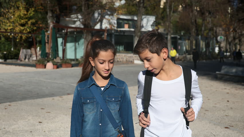 Steadicam shot of cute pretty kids, a boy and a girl with school bags walking together in the city to go to school in spring or summer. Friends classmates or brothers.