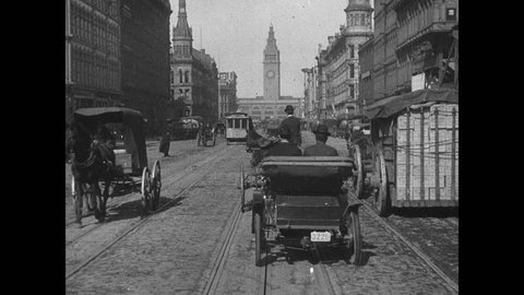 1900s: Cars and horse drawn carriages travel down city street. Cable car travels along tracks down street. People walk around city.