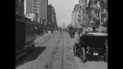 1900s: Cars and horse drawn carriages travel down city street. Cable cars travel along tracks down street. People walk around city. Man rides bicycle, men stand in street.