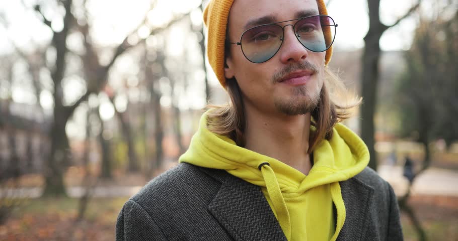 4k. Autumn vibes. Handsome hipster in the park. Young man with beard in yellow heat walks across the park and looks smilingstraight in the camera