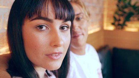 Millennial girl stares at camera while her boyfriend has his arm around her in cafe on wood bench in Australia during the day. Medium to closeup shot with 4K RED camera.