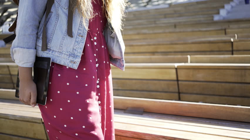 Faceless shot of woman in denim and dress holding tablet and walking in modern park