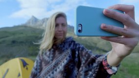 attractive girl hiking makes selfie on smartphone on a background of mountains. A girl stands near the tent, camping with a tent. The concept of freedom in adventures in mountains. Wearing a poncho.